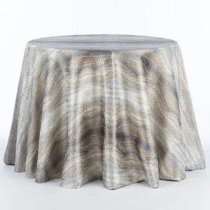 A round table is adorned with a flowing, patterned Magma Mixed Metal tablecloth featuring shades of grey, beige, and white, available for rental.