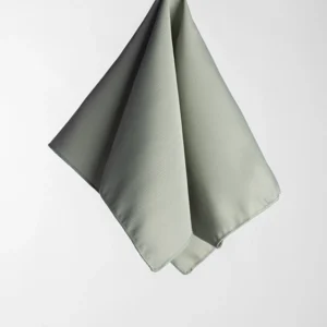 The Monaco Seafoam fabric napkin, featuring a subtle texture and light gray hue, hangs folded against a plain white background.