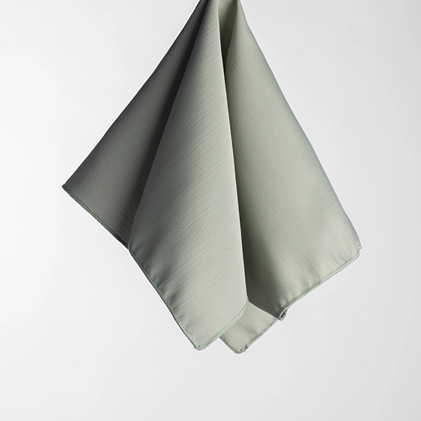 The Monaco Seafoam fabric napkin, featuring a subtle texture and light gray hue, hangs folded against a plain white background.