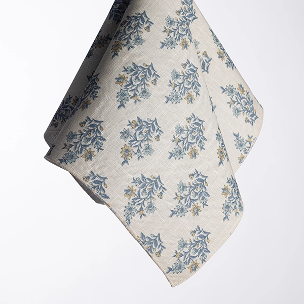 A piece of Morgan Natural fabric with a grey background and a repeating pattern of blue and yellow floral designs draped in mid-air against a plain white backdrop.