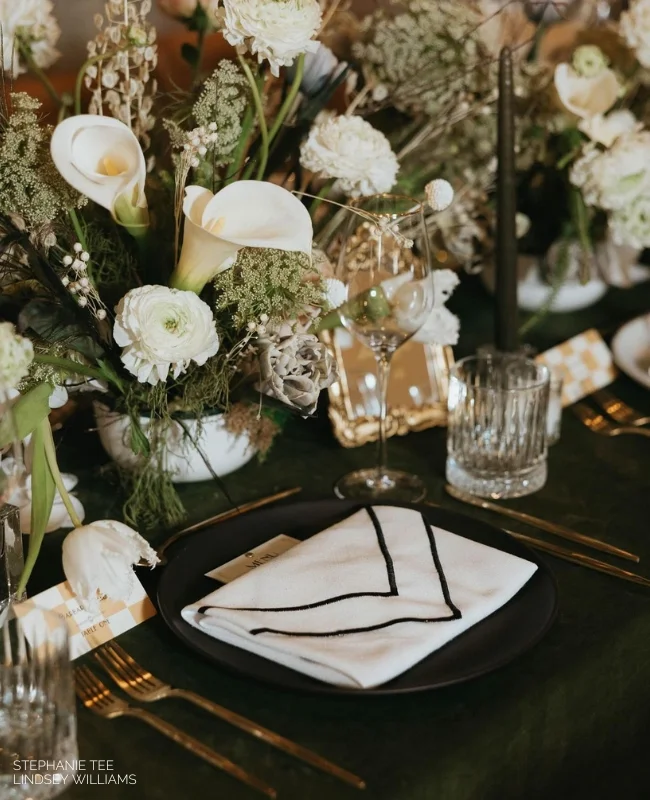 Elegant dining table setup featuring a black plate with sophisticated napkin folding ideas for weddings, surrounded by glasses and white floral arrangements.