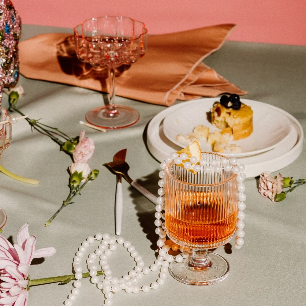 A table setting with a glass of orange drink, flowers, a piece of cake on a plate, and a beaded accessory. Pink napkins are folded in the background.