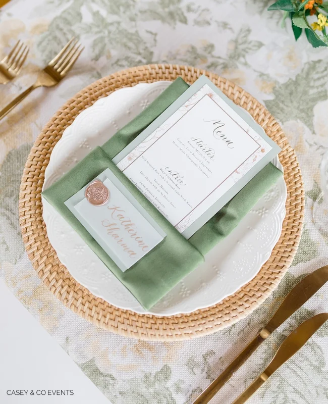 Elegant table setting featuring a woven placemat with a white plate, sophisticated napkin folding, and a menu card sealed with a wax stamp, complemented by gold flatware.