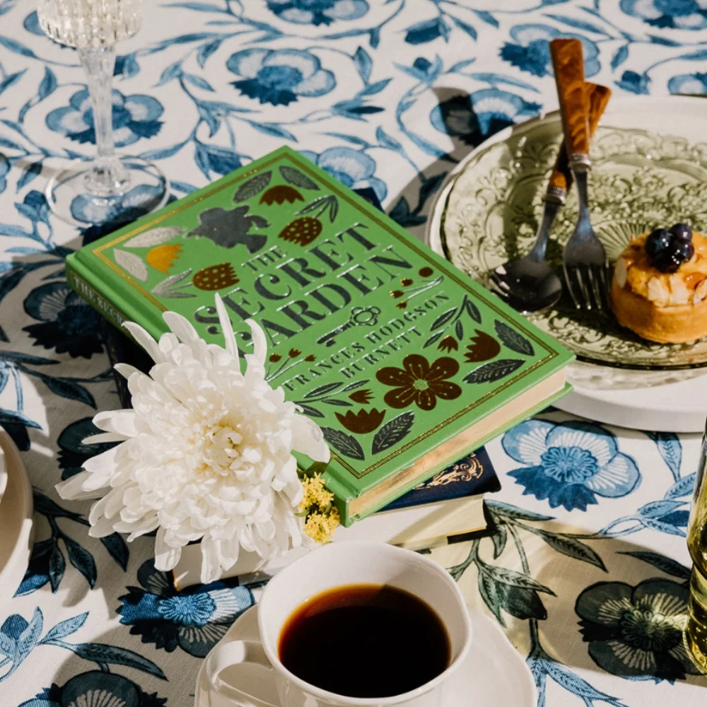 A neatly laid table with a floral-patterned tablecloth features a book titled "The Secret Garden," a dessert plate with a pastry, a cup of coffee, a white flower, and silverware.