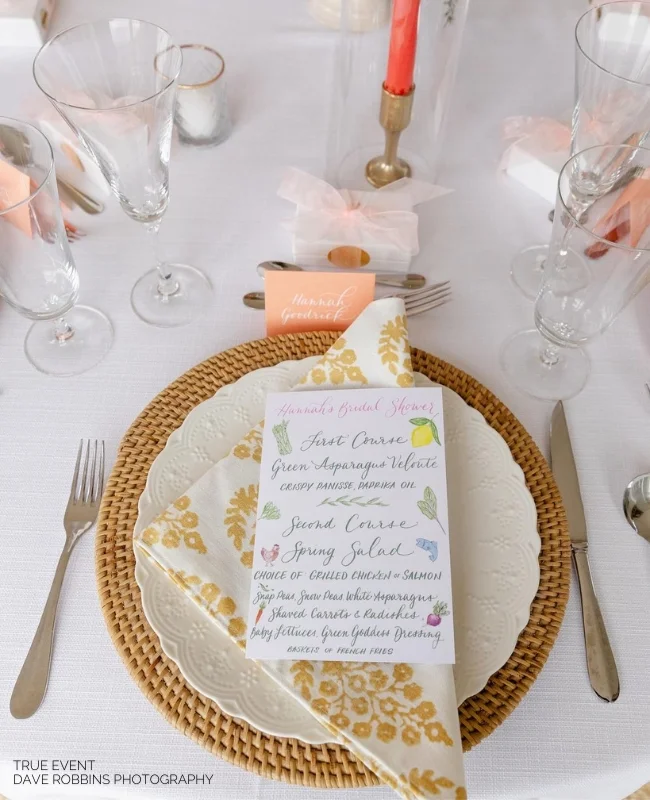 Wedding table setting with detailed event napkin folds on a gold-patterned napkin, circular wicker placemat, and elegant tableware, featuring soft pink accents and candles.