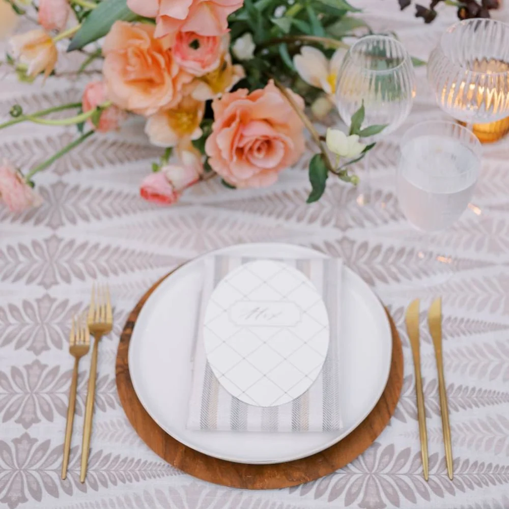Stylishly set table with floral arrangement, white plate on wooden charger, gold utensils, and glasses. A striped napkin with an oval card rests on the plate.