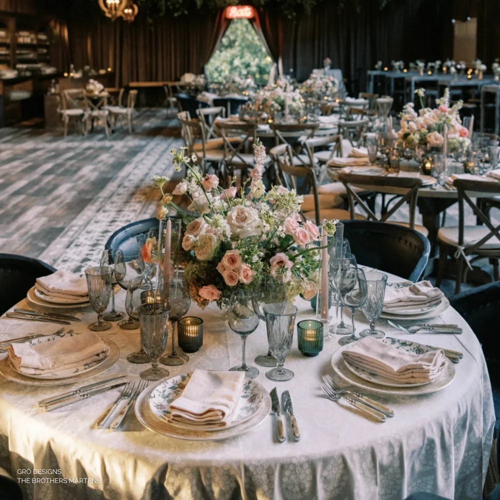 A round table set for a formal event with floral arrangements, candles, and glassware. More tables similarly arranged are in the background.