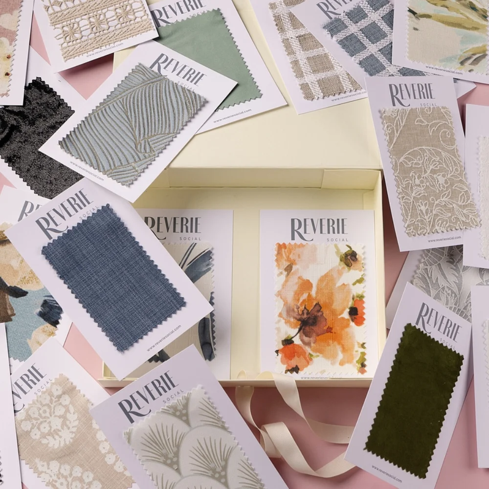 A collection of fabric swatches in various colors and patterns surrounds an open box holding two swatches with "Reverie" branding. Each swatch is attached to a card with pink background visible below.