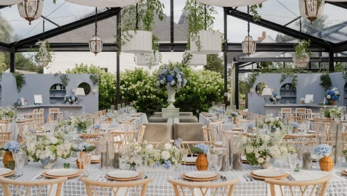 A beautifully decorated indoor venue for a wedding reception, featuring elegant floral arrangements, checkered tablecloths from our linen rentals, wooden chairs, overhead lanterns, and a glass ceiling with hanging greenery.