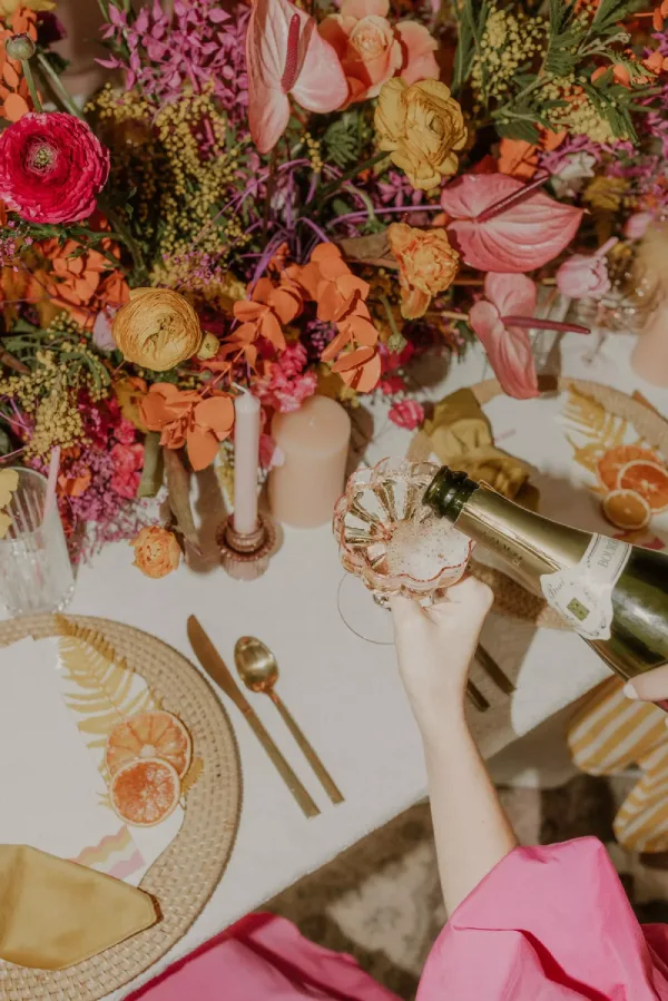 A person pours champagne into a glass on a table set with vibrant flowers, a small candle, plates adorned with orange slices, gold-tone cutlery, and elegant linens from our table linen rental collection.