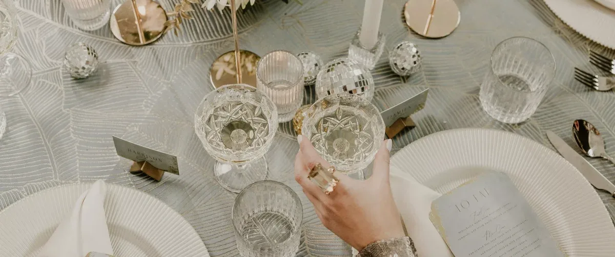 A hand holding a crystal champagne glass at an elegantly set dining table with white plates, glasses, and decorative elements, including candlesticks and small disco balls. The immaculate presentation is elevated by exquisite linen rentals that add a touch of sophistication to the setting.