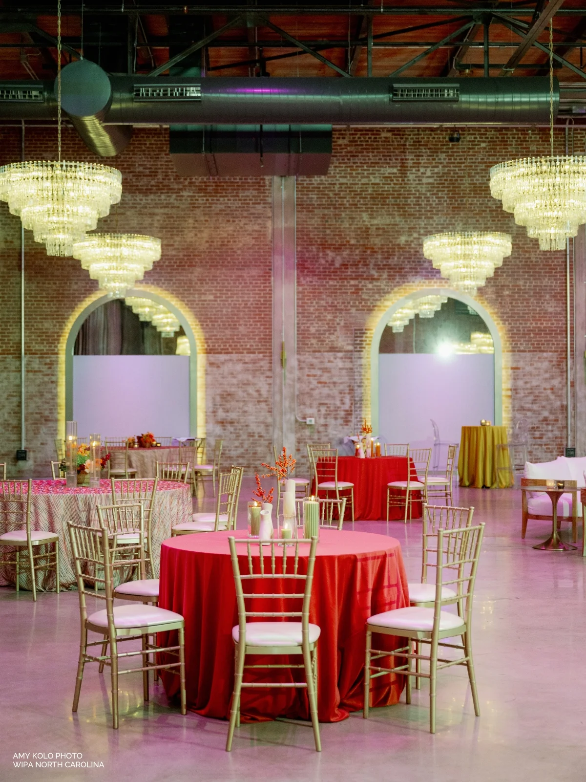A decorated event space features round tables with red tablecloths, gold chairs, and elegant chandeliers hanging from the ceiling. Brick walls with large arched mirrors are in the background.