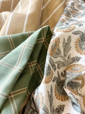 Close-up of three folded fabrics with different patterns: a brown fabric with white stripes, a green fabric with white checks, and a white fabric with a floral and leaf design.