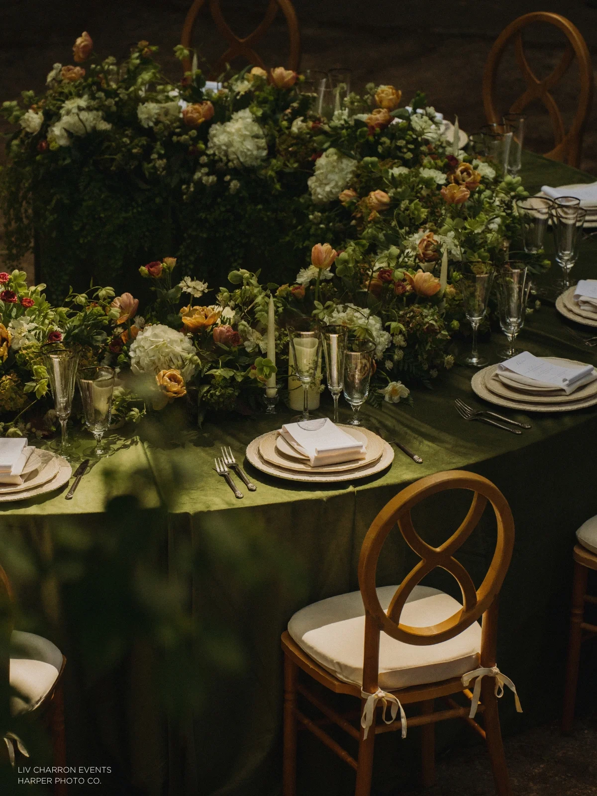 A circular dinner table is decorated with green foliage and flowers, set with plates, glasses, and white napkins. Wooden chairs with white cushions are arranged around the table.