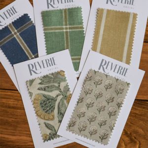 Introducing "The Swatch Edit Vol. 2": a collection of six fabric swatches on cards branded with "Reverie Social." The fabrics showcase diverse patterns, including plaid and florals, in an array of green, blue, and tan hues, all beautifully displayed on a wooden surface.