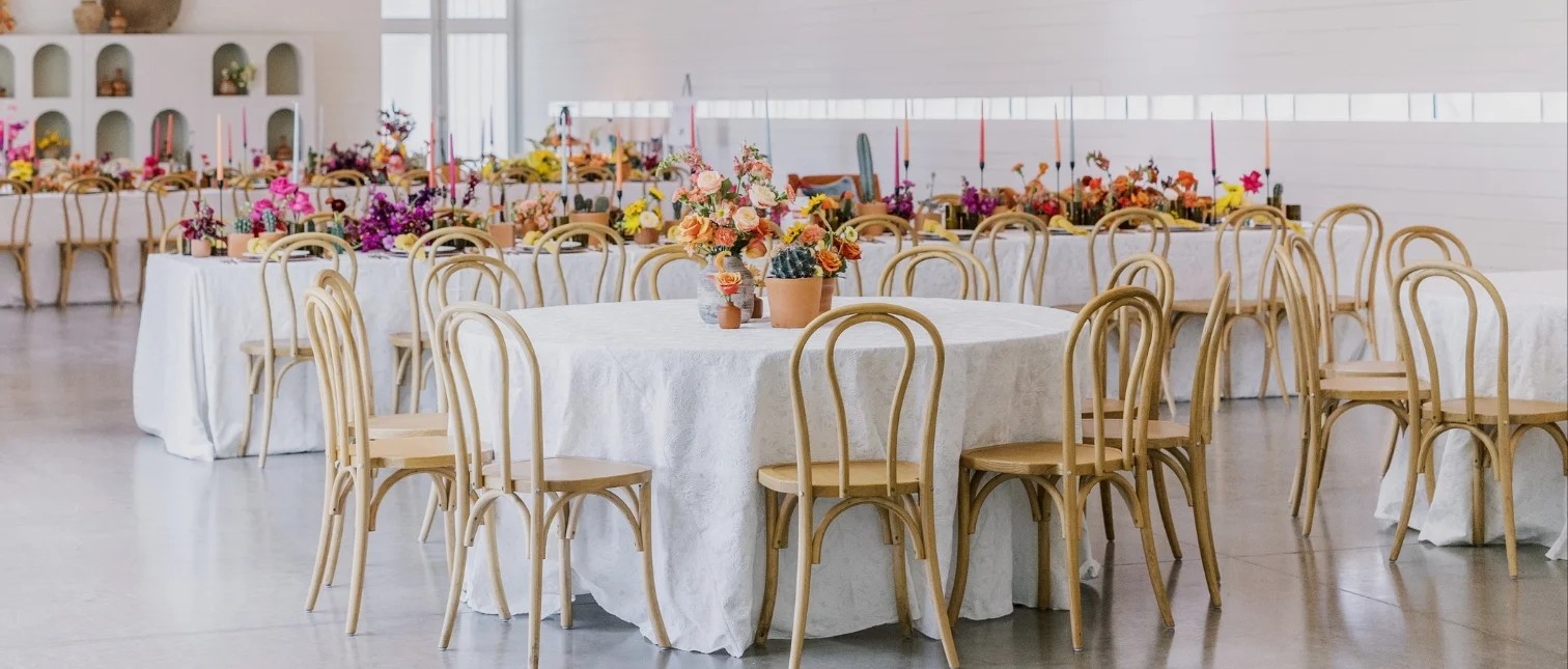 A decorated dining area features round tables with white tablecloths, wooden chairs, and colorful flower centerpieces, set in a bright room with large windows.