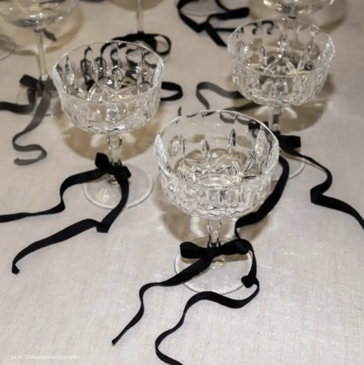 Three empty crystal champagne coupes with black ribbon bows tied around the stems are arranged on a white tablecloth.