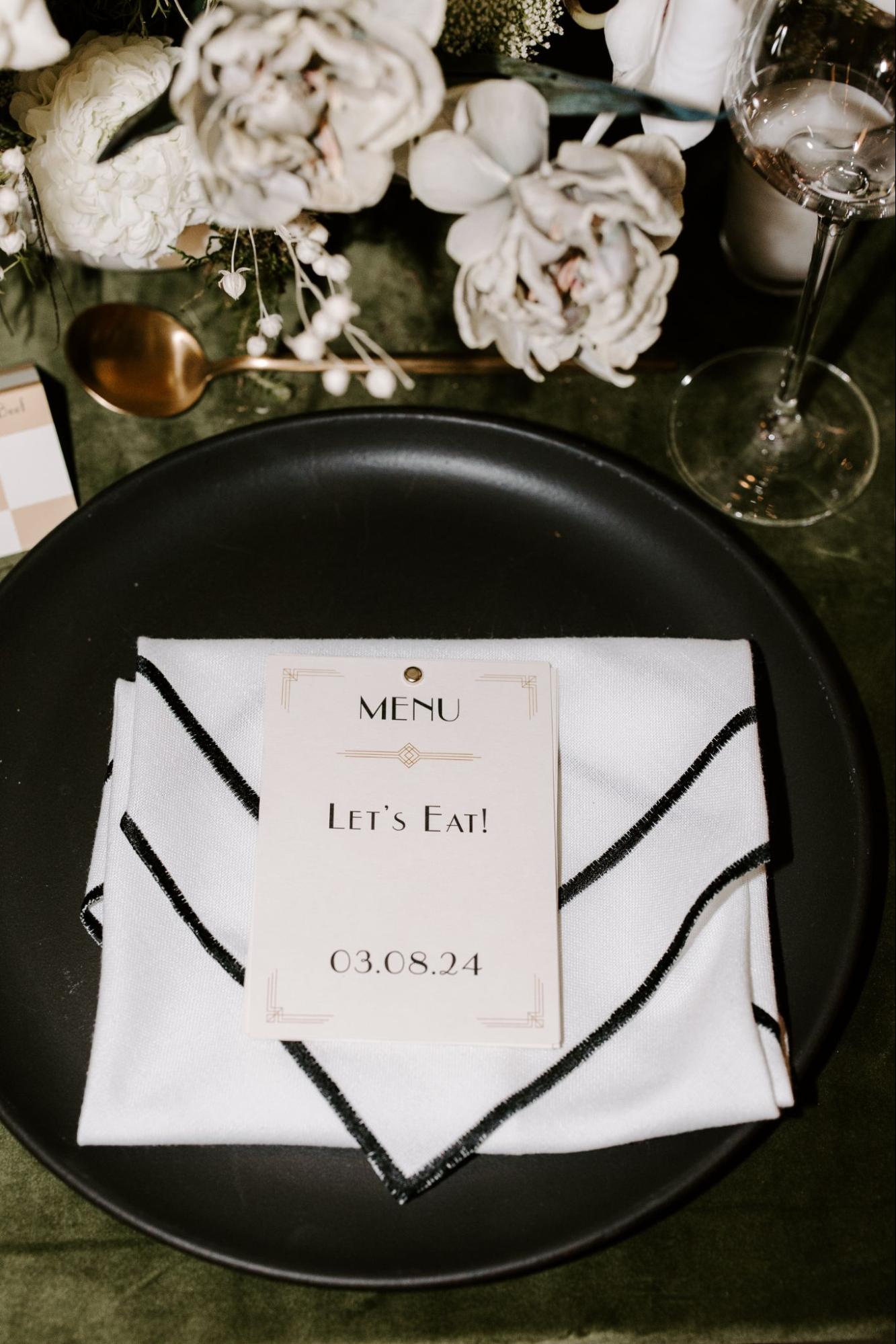 A plate with a folded white napkin is placed on a dark green tablecloth, with a menu card on top reading "Let's Eat! 03.08.24." Flowers and a wine glass are next to the plate.