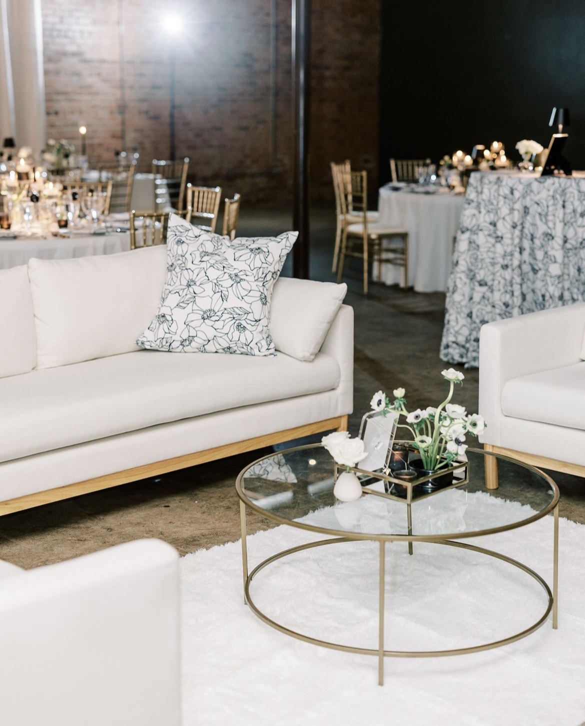 A modern lounge area with white sofas, a clear glass coffee table, and floral decor. Tables and chairs are set up in the background for a formal event, with soft lighting enhancing the ambiance.