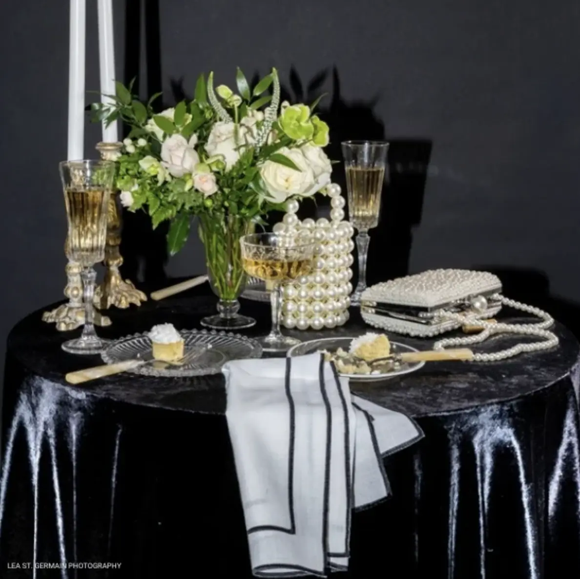 A table with elegant decorations including a bouquet of flowers, champagne glasses, a pearl handbag, a pearl-covered clutch, and a black velvet tablecloth with plates of unfinished food.