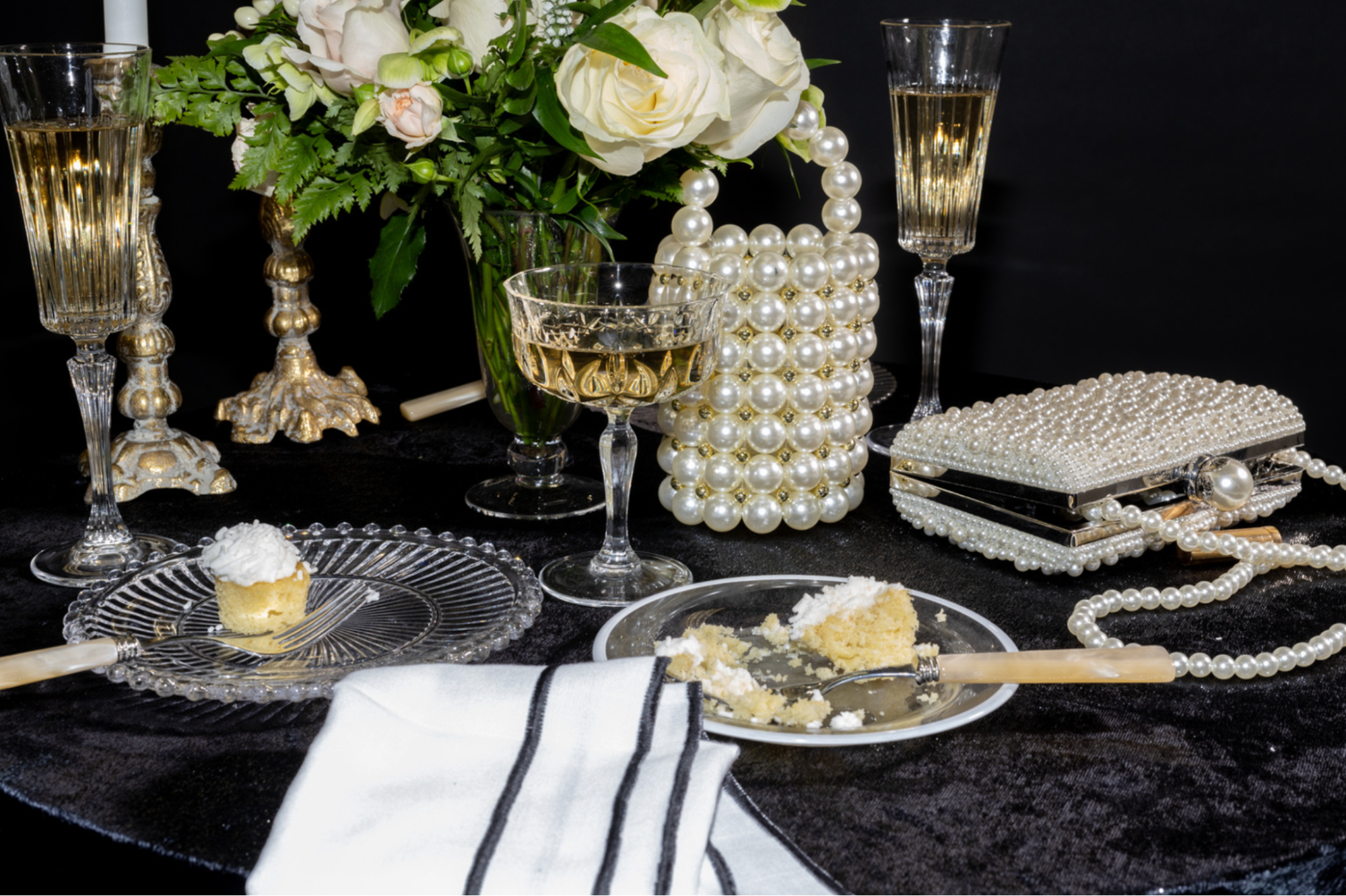 A table setting featuring a partially eaten slice of cake on a plate, cake crumbs, champagne glasses, a floral arrangement, and pearl-studded handbags against a black background.