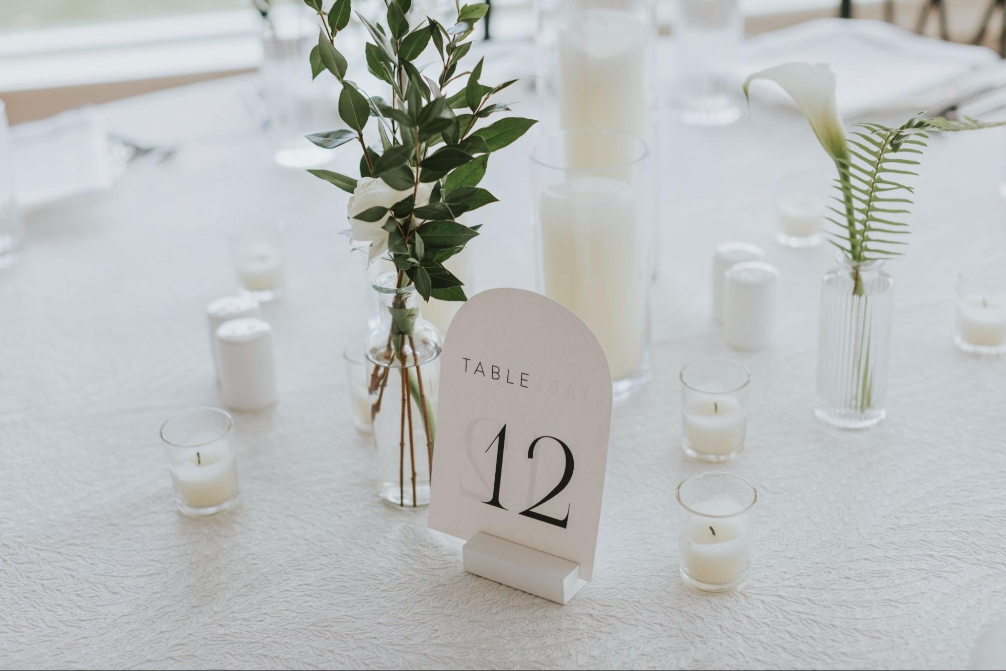 A table with white flowers in vases, candles, and a sign that reads "Table 12" against a textured white tablecloth.