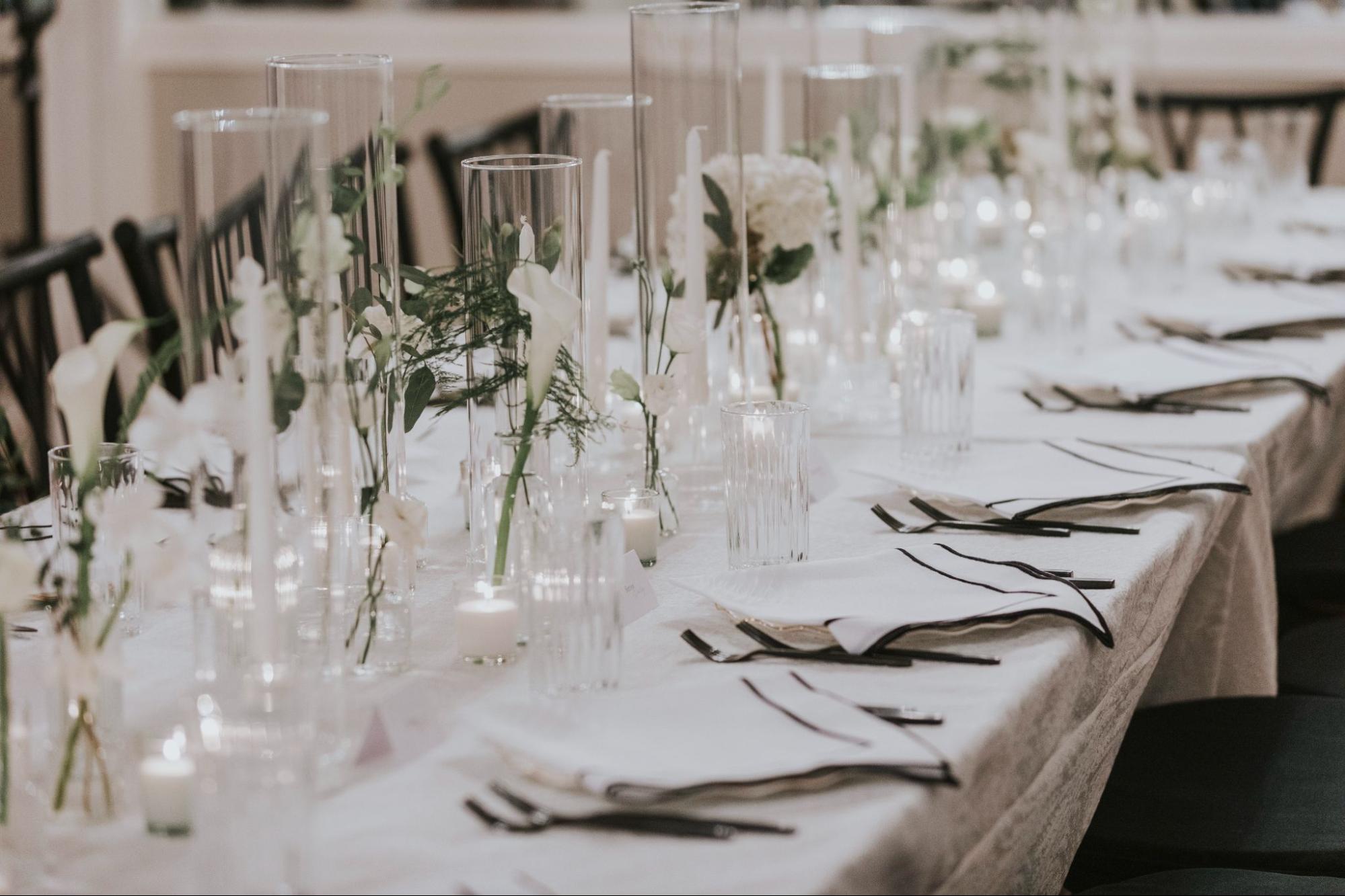 A long table is set with white linens, black chairs, glassware, white flowers, candles, and tall glass vases.