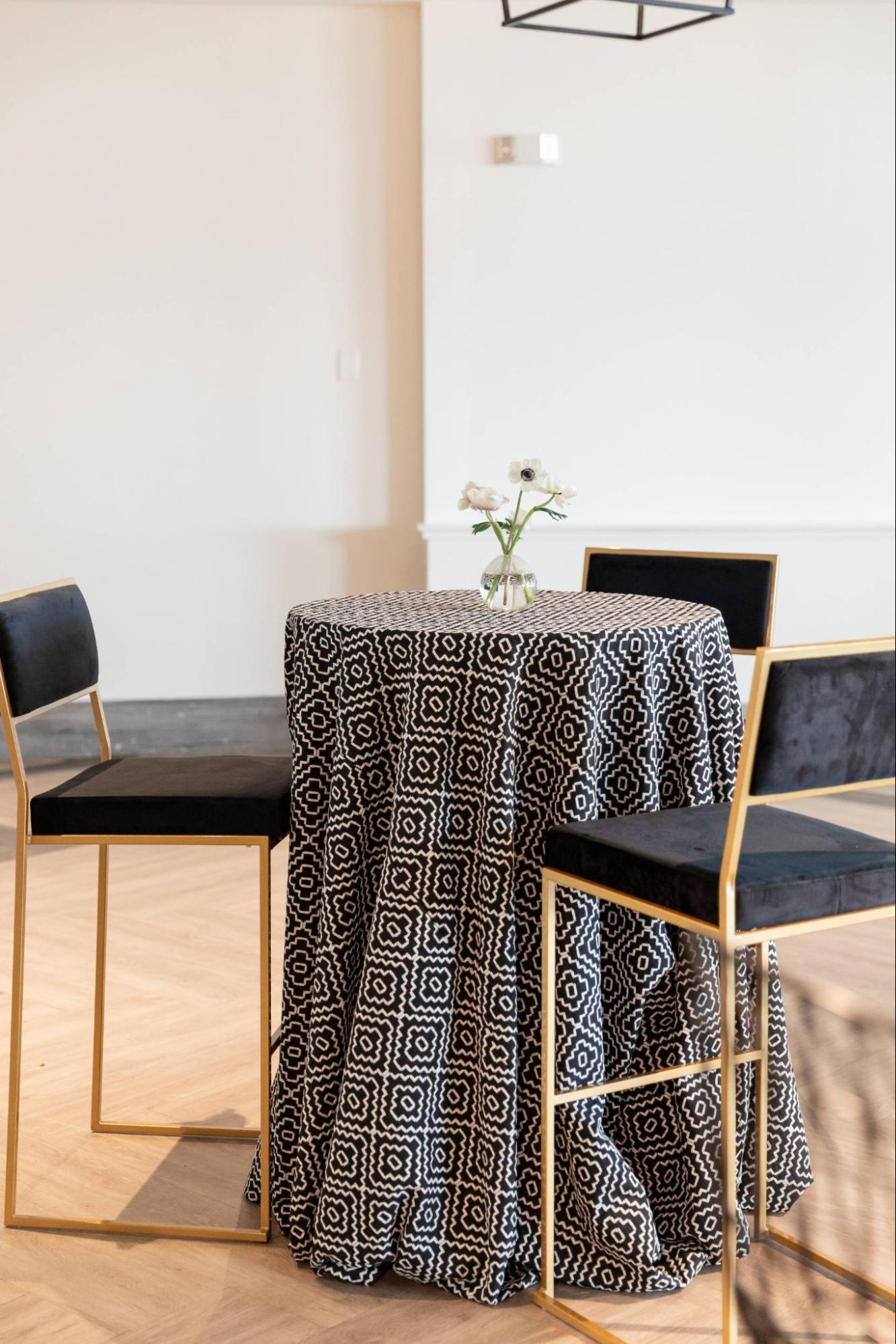 A small round table with a black and white patterned tablecloth, a vase of white flowers on top, and two black chairs with gold frames.