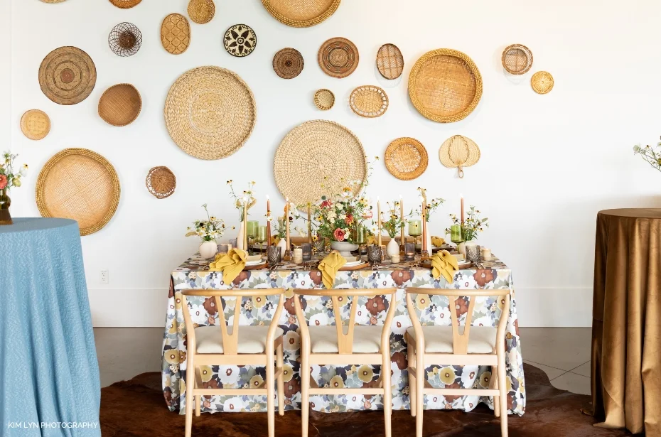 A dining table set with floral tablecloth, tan napkins, floral centerpieces, and candles. The wall behind it is decorated with various woven baskets. Three chairs are at the table.