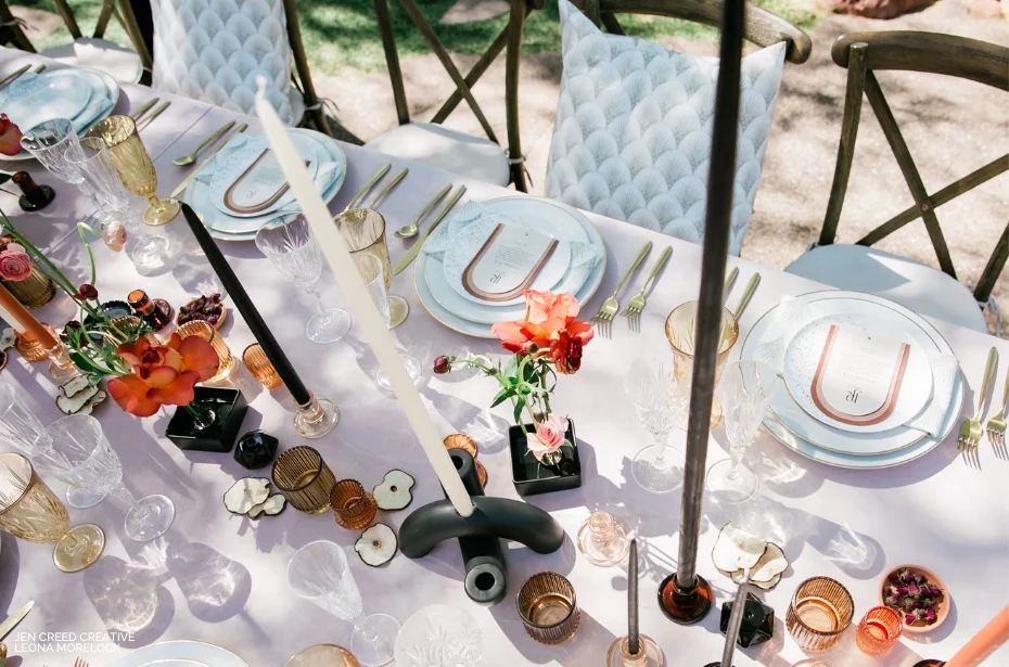 A top view of an elegant outdoor dining table set for a meal with white plates, gold utensils, black candles, and flower arrangements. The tablecloth is light pink, and seating includes cushioned chairs.