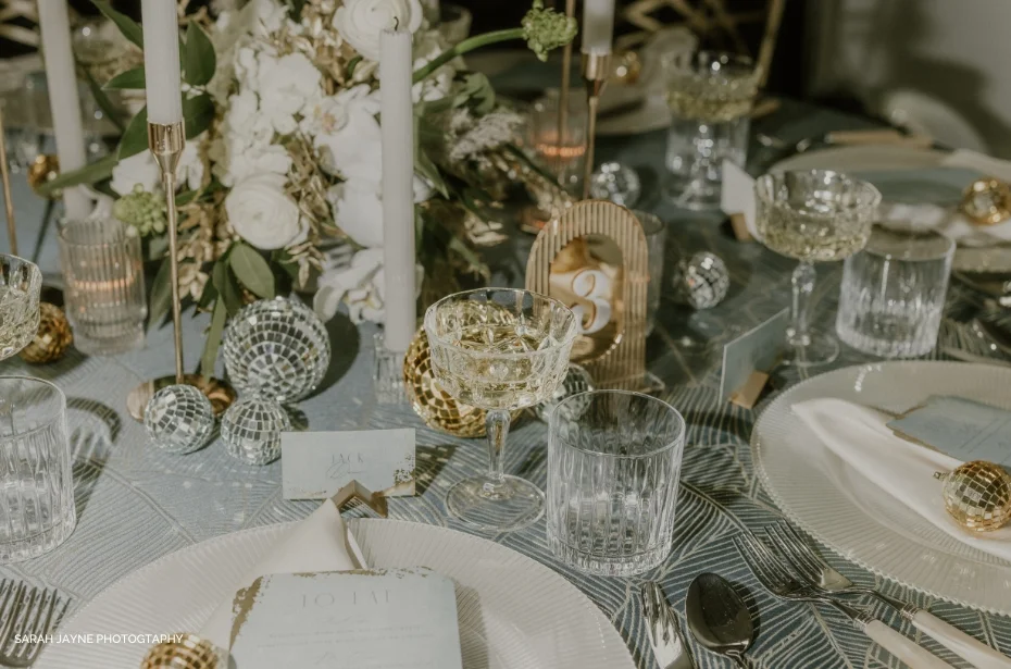 A formal table setting with white plates, crystal glasses, floral centerpieces, gold table numbers, candles, and decorative ornaments.