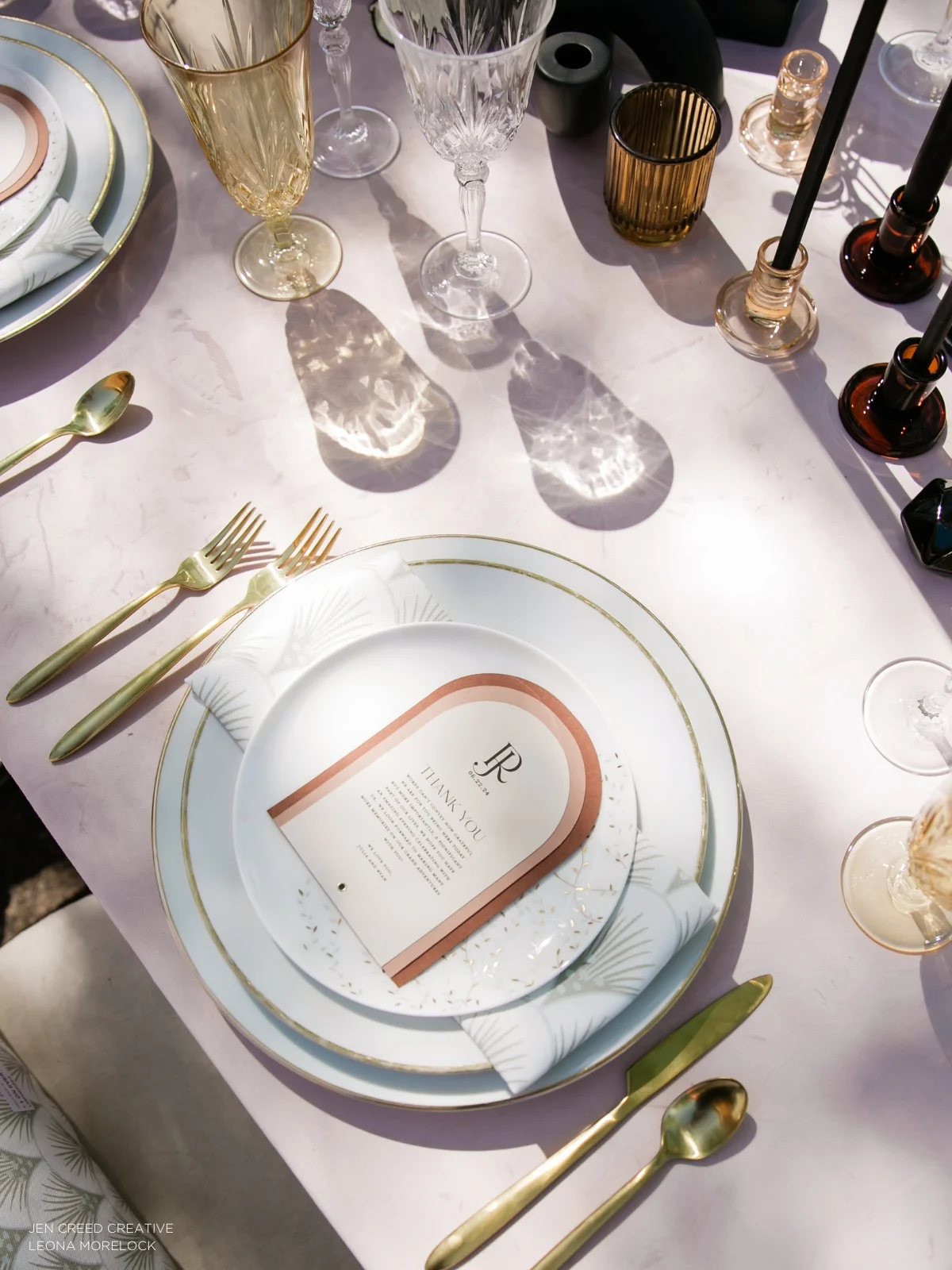 An elegantly set dining table with gold-trimmed plates, gold utensils, various glassware, and a thank-you card at each place setting. Candles and decorative items are also visible on the table.