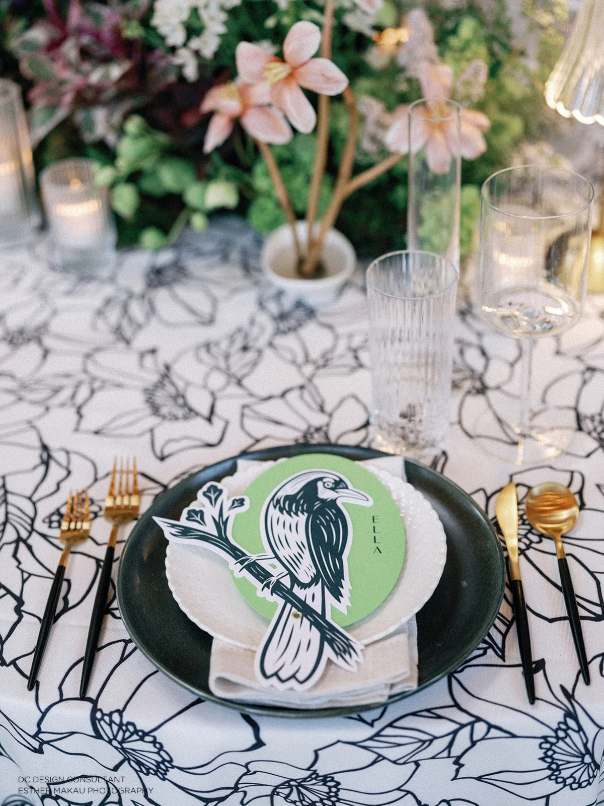 A place setting with a black and white bird illustration on a green plate, atop a floral tablecloth with glasses and cutlery arranged. Flowers and candles are in the background.