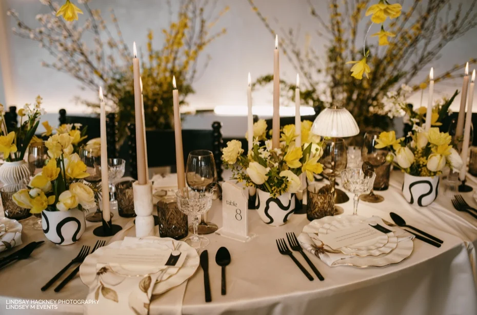 A table set for a formal event, featuring white and black patterned tableware, black cutlery, white napkins, yellow floral arrangements, and tall candles.