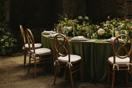 A round table set for a meal with green tablecloth from our table linen rental collection, floral centerpiece, and white cushioned chairs in an outdoor setting.