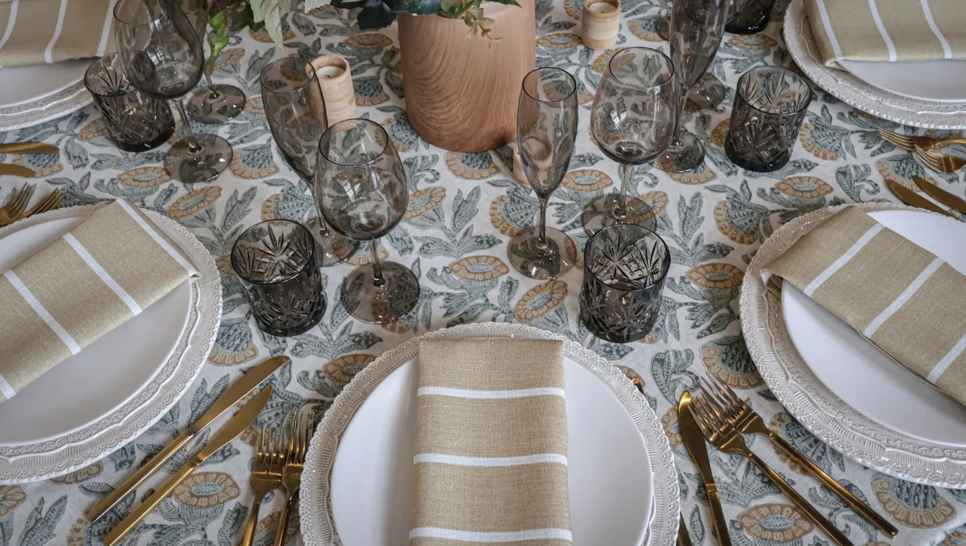 A table set for a meal with patterned tablecloth, white plates, striped beige napkins, gold cutlery, and various glassware, including water and wine glasses. The centerpiece features greenery. Consider table linen rental for an effortlessly elegant setting.