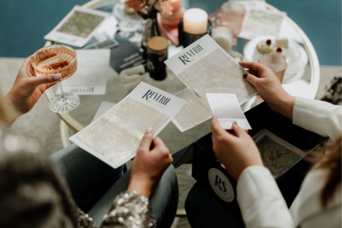 Two people sit at a round table reviewing textured paper samples and holding drinks, with various items including candles, desserts, and brochures for table linen rental on the table.