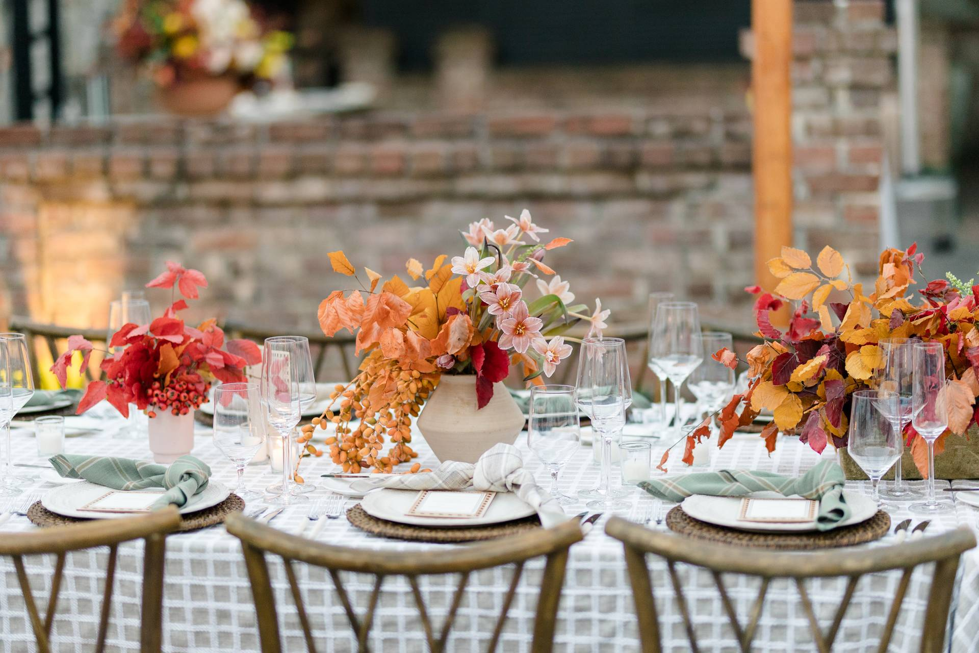 A table set for a meal, featuring fall-themed floral arrangements, plates, glasses, and cutlery, with wooden chairs arranged around it.