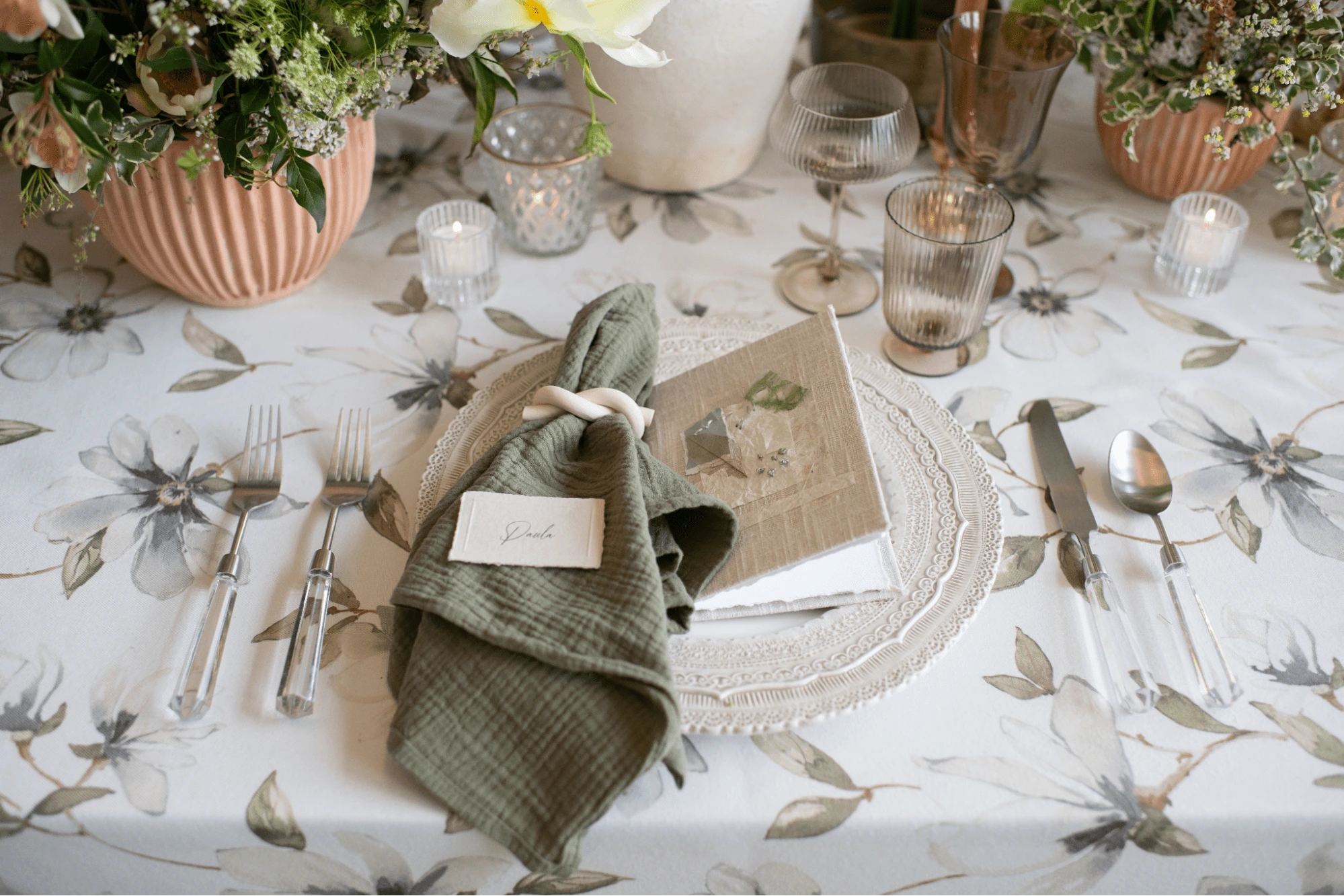 A decorated table setting with floral tablecloth, glassware, candles, and a ceramic planter with flowers. The place setting includes a napkin with a name card, utensils, and a gift box on top of a plate.