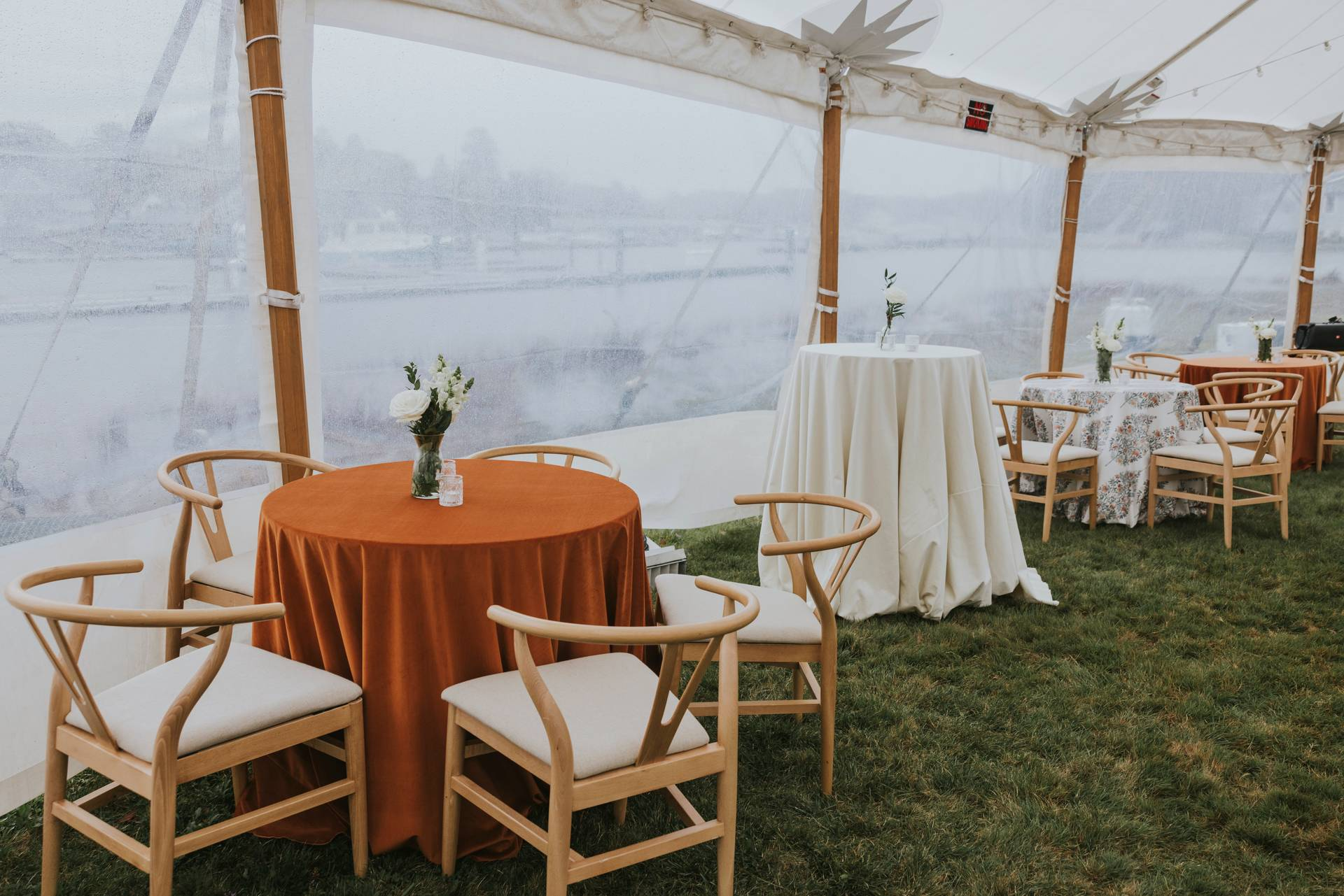 A tented event space features round tables with various fabric covers and wooden chairs, set atop a grassy surface. The clear tent walls reveal a foggy, outdoor background with indiscernible scenery.