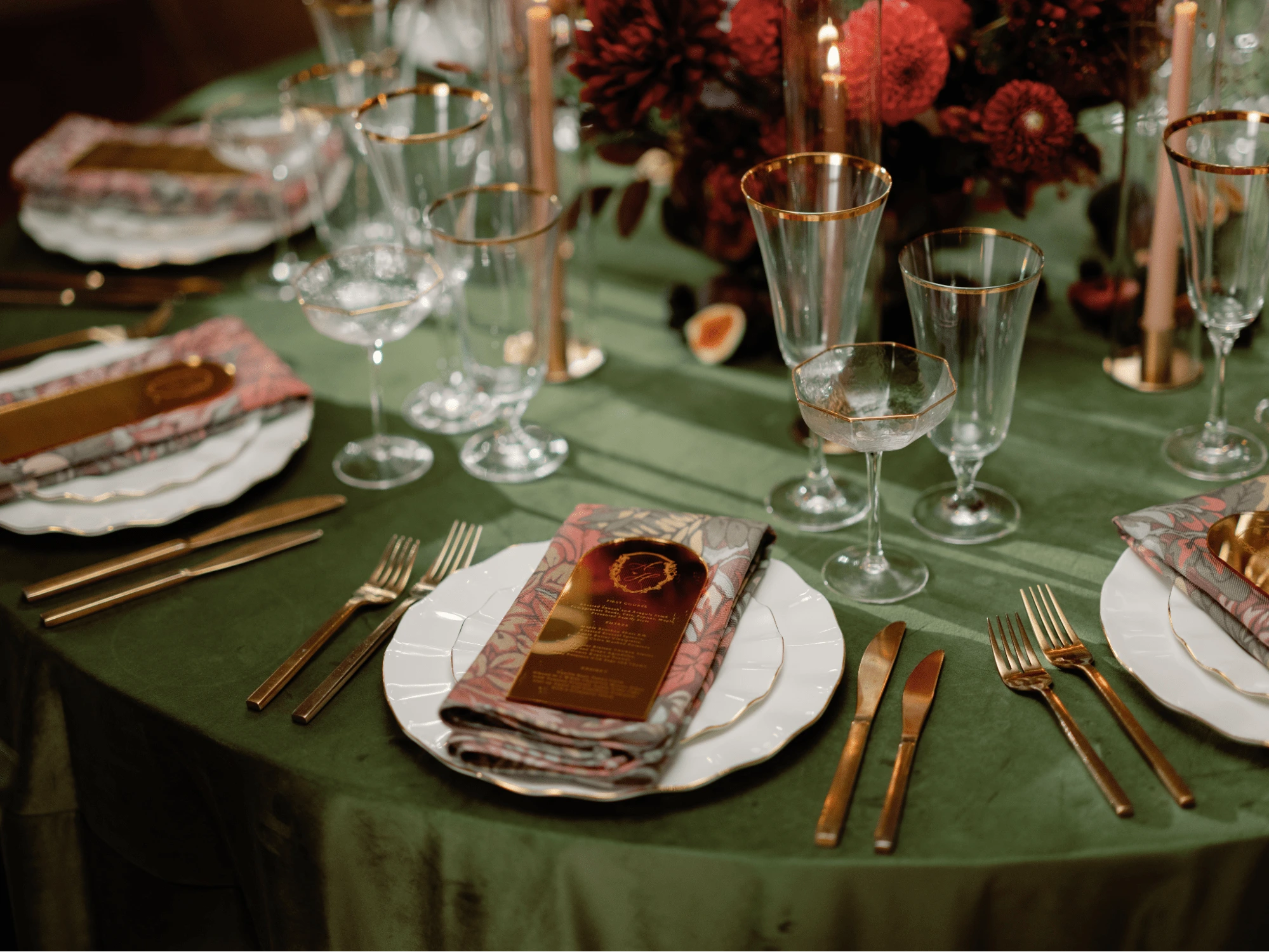 A table set for a formal dinner with green tablecloth, white plates, folded napkins, gold cutlery, various glasses, and floral centerpieces.