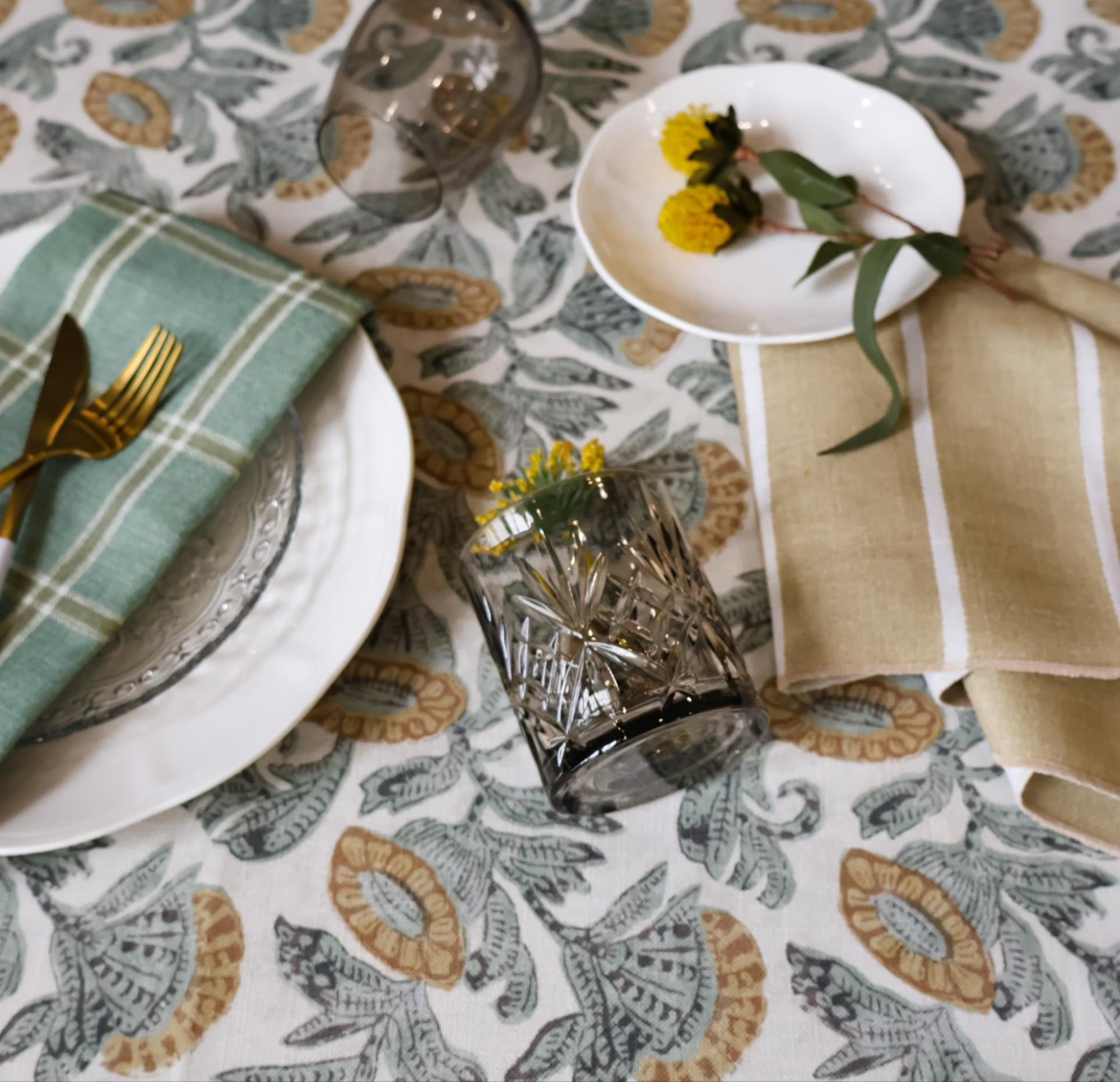 A dining table setup with floral tablecloth, a green plaid napkin, gold cutlery, a glass with yellow flowers, and a small plate with yellow blooms on a beige napkin.