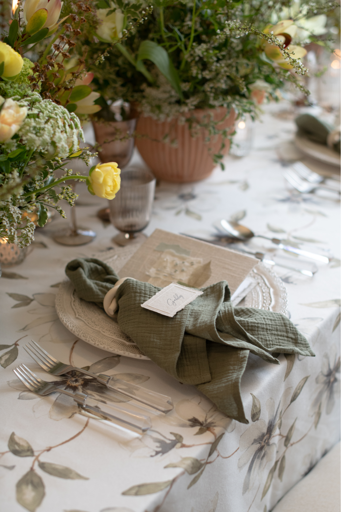 A table is set with green napkins, white plates, silver cutlery, and name cards. Floral arrangements and candles are placed as centerpieces on the floral-patterned tablecloth.