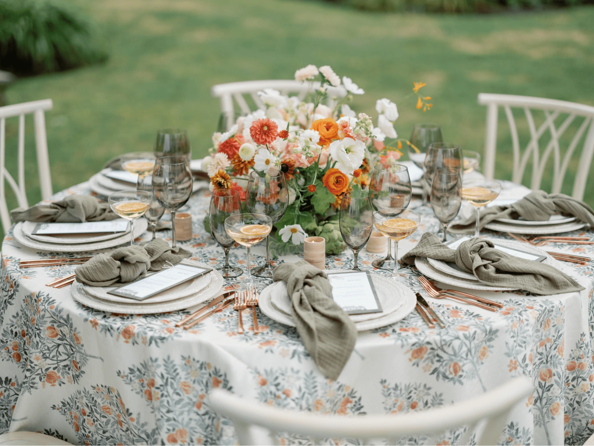 A round table set for a meal with floral tablecloth, neatly arranged tableware, gray napkins, and a central bouquet of colorful flowers. Six chairs surround the table, which is situated on a lawn.