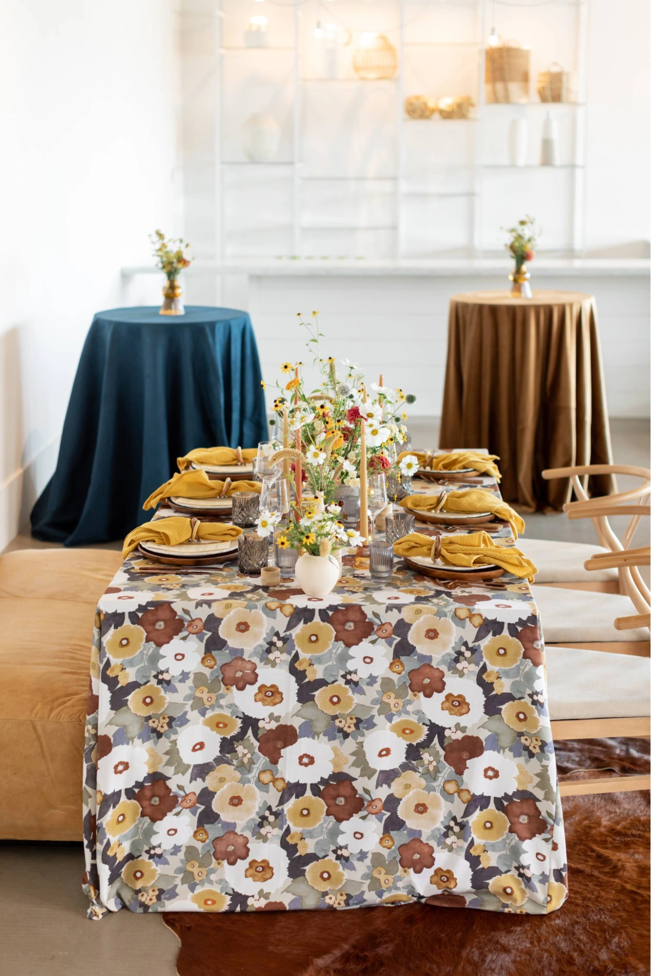 A dining table with a floral tablecloth, yellow napkins, and small flower arrangements. Background: two round tables with blue and brown tablecloths and additional flower arrangements.