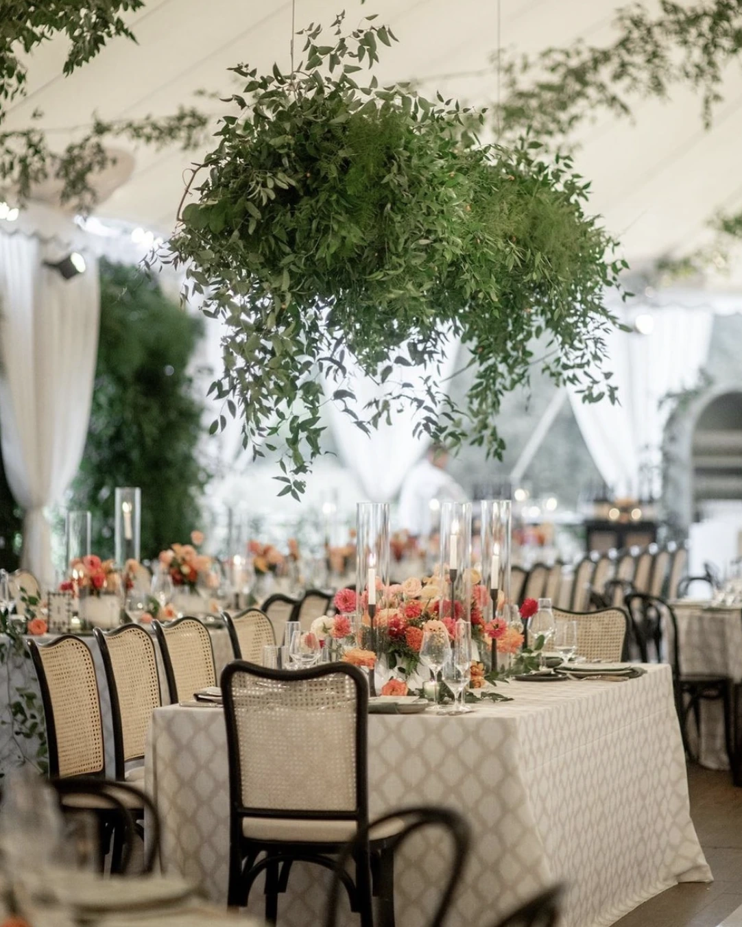 Elegant dining setup with white tablecloths, floral centerpieces, and hanging greenery under a tent. Rows of chairs surround the tables, set for a formal event.