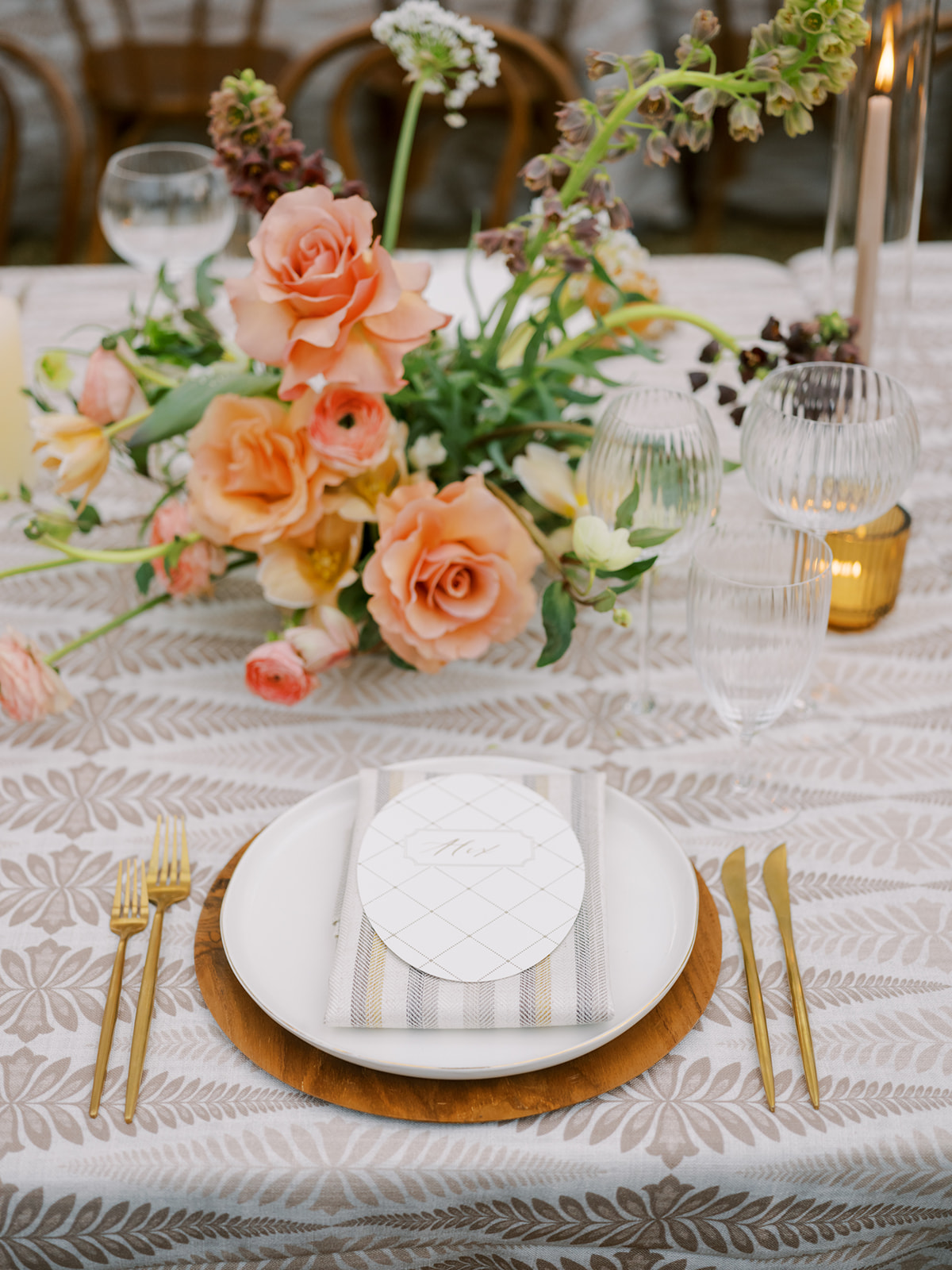 A dining table is set with a patterned tablecloth, a white plate, gold cutlery, a menu card, and an arrangement of peach and pink flowers. Glassware and candles are also arranged on the table.