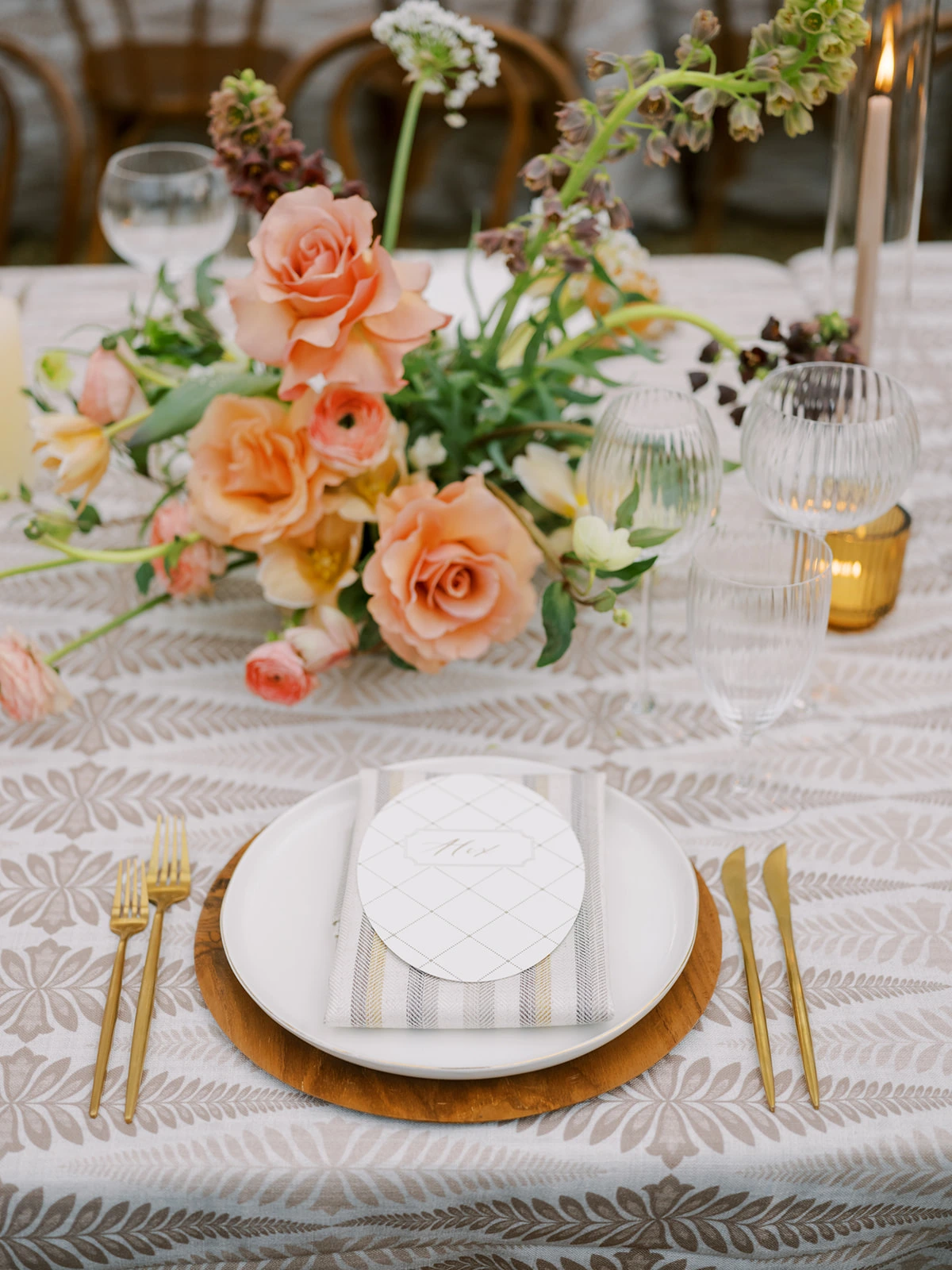 A dining table is set with a patterned tablecloth, a white plate, gold cutlery, a menu card, and an arrangement of peach and pink flowers. Glassware and candles are also arranged on the table.