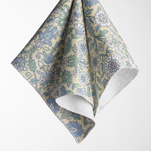 A square piece of fabric with a floral pattern in blue, green, and white hues against a light background, with one corner folded over, showing a white underside.