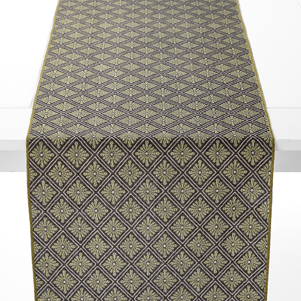 A table runner with a diamond and floral pattern in shades of gold and black is draped over a white table.